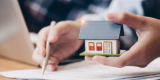 One hand holding a house model and one hand holding a pen going through documents